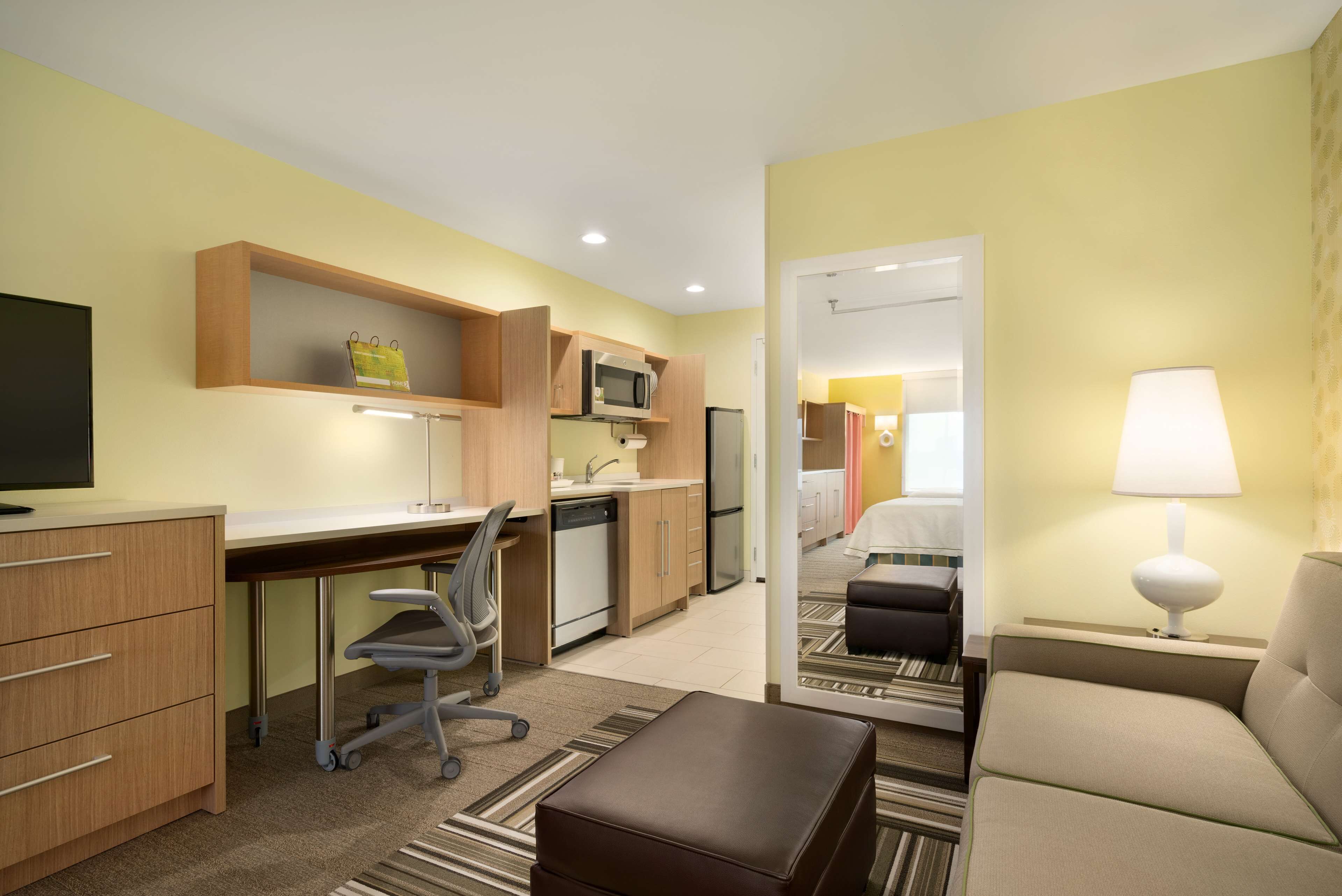 A compact hotel room with a kitchenette, a desk, a chair, a sofa, and a TV.
