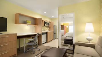 A compact hotel room with a kitchenette, a desk, a chair, a sofa, and a TV.