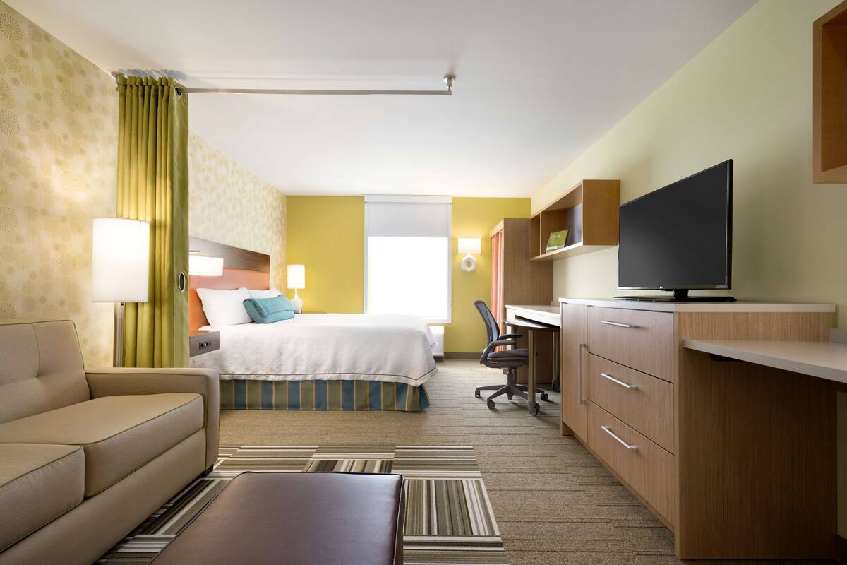 Une chambre d’hôtel avec un lit, un bureau équipé d’un ordinateur, une télévision, un canapé et une salle de bain visible à travers une porte ouverte.