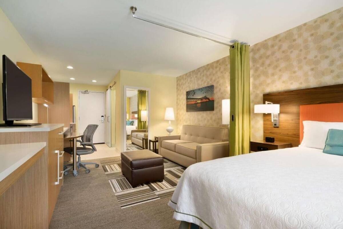 Une chambre d’hôtel avec un lit, un bureau, une chaise, un pouf et un tableau au mur.