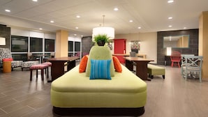 A modern hotel lobby with a large green sofa, a potted plant, and a dining area with red chairs.