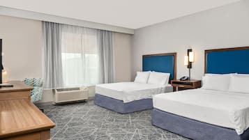 A hotel room with two beds, a desk, a chair, a window with sheer curtains, an air conditioning unit, and a wall-mounted phone.