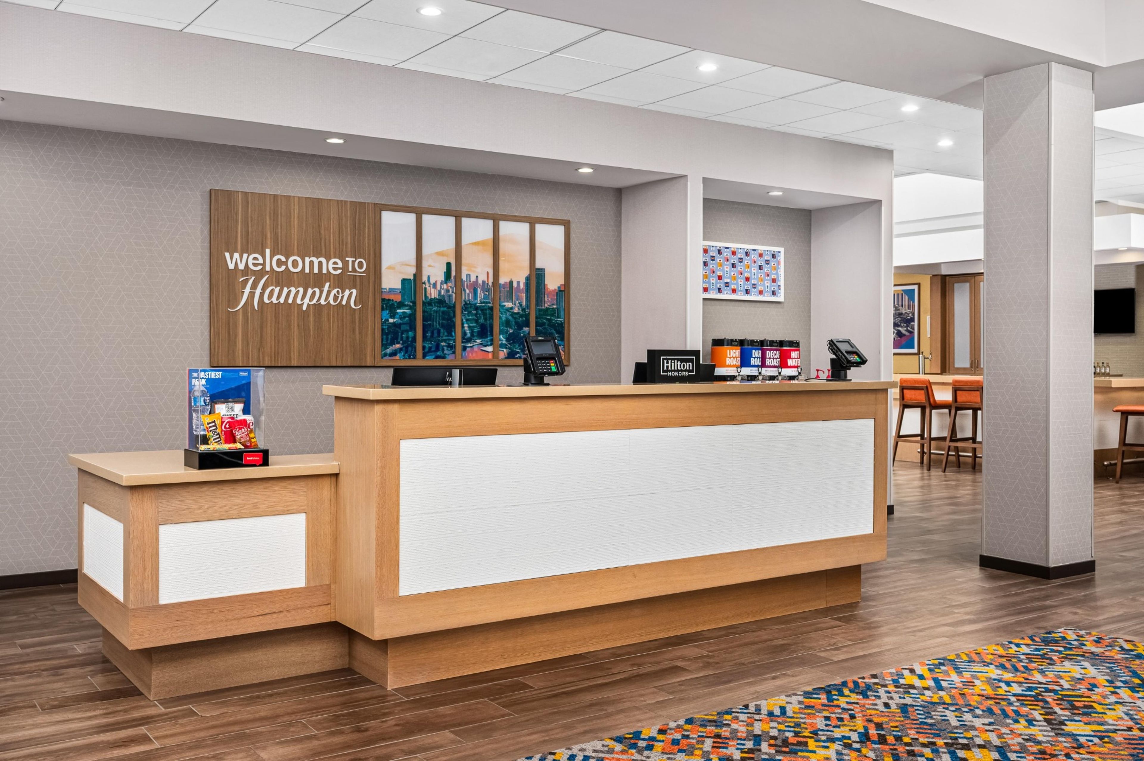 A hotel reception desk with a 'Welcome to Hampton' sign, a digital kiosk, and a seating area with a patterned rug.