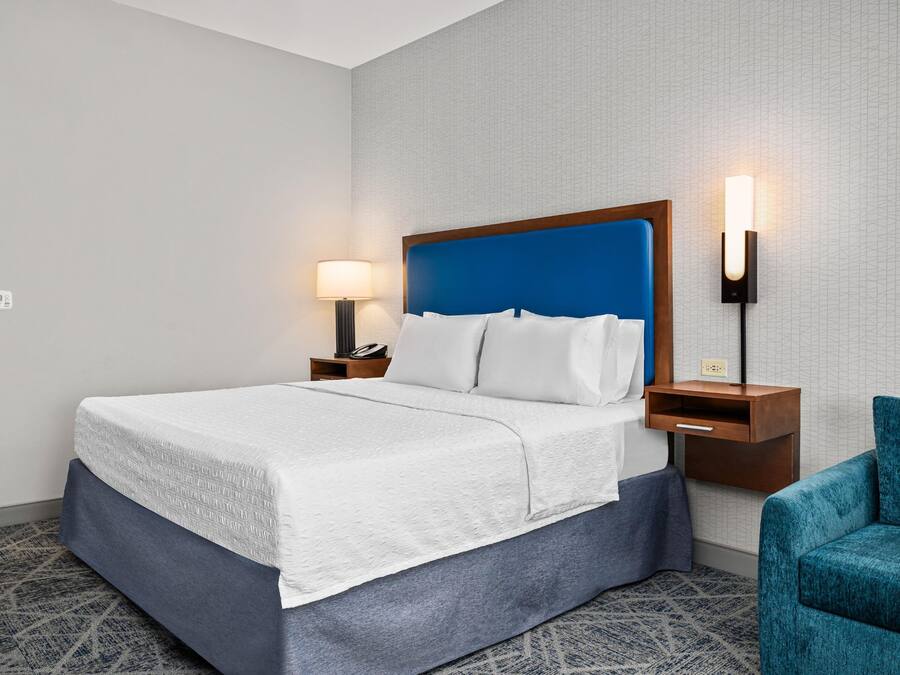 A hotel room with a bed, a nightstand, a lamp, a chair, and a wall-mounted light fixture.