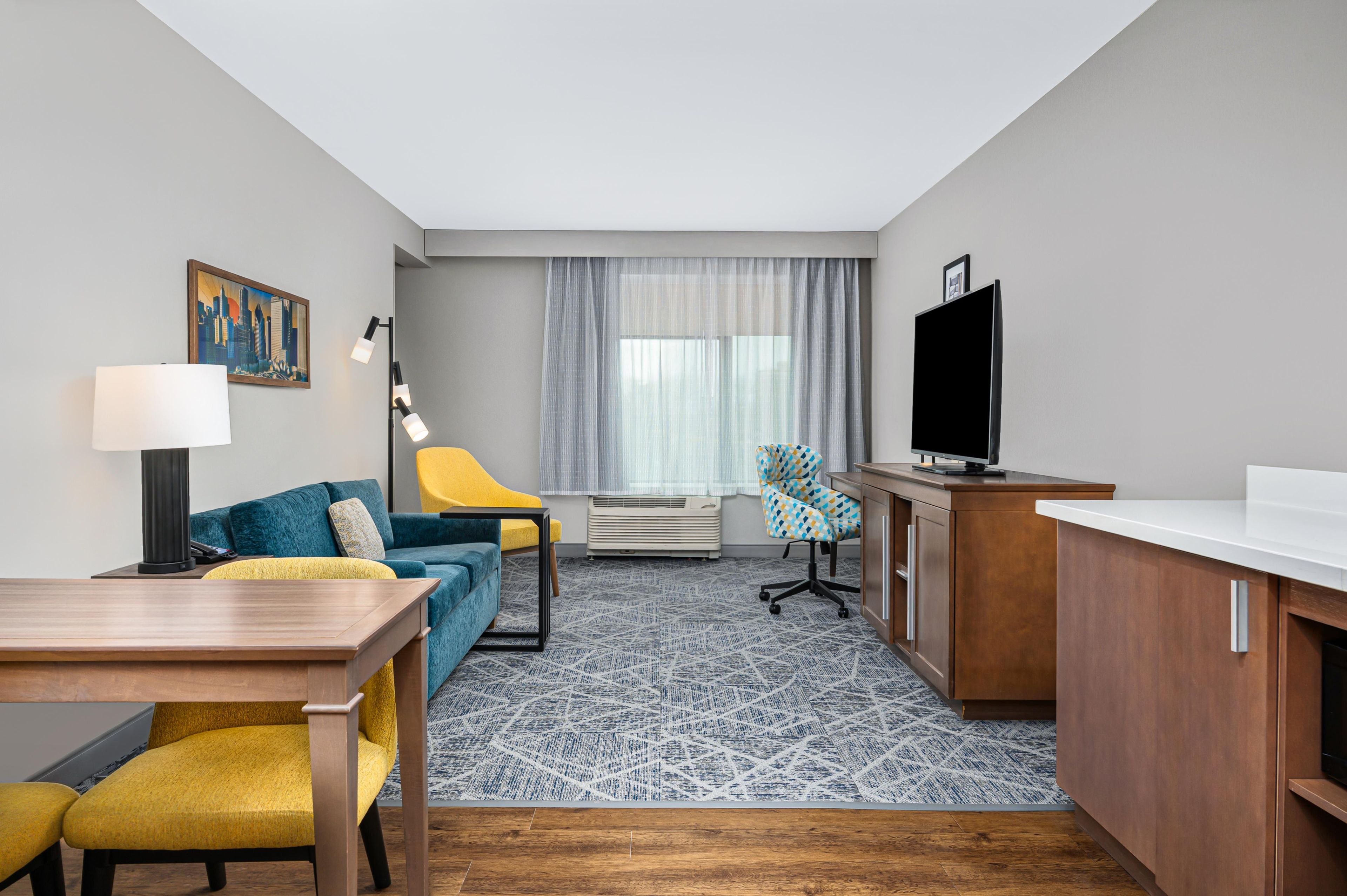 Habitación de hotel compacta con sofá, una silla amarilla, escritorio con lámpara, televisor y alfombra estampada.