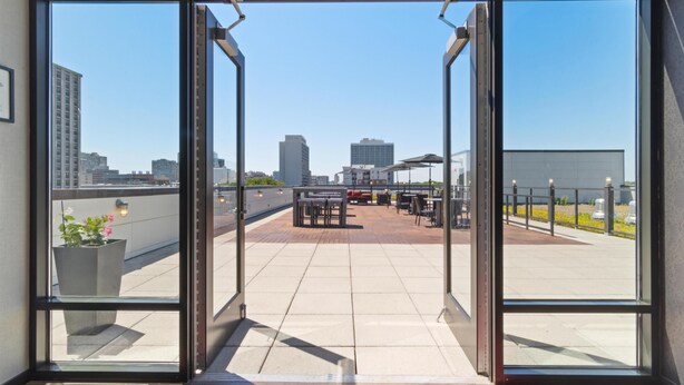 A view through a glass door to an outdoor patio area with tables and chairs, a potted plant, and a clear sky. - Hampton Inn Chicago North-Loyola Station (Chicago)