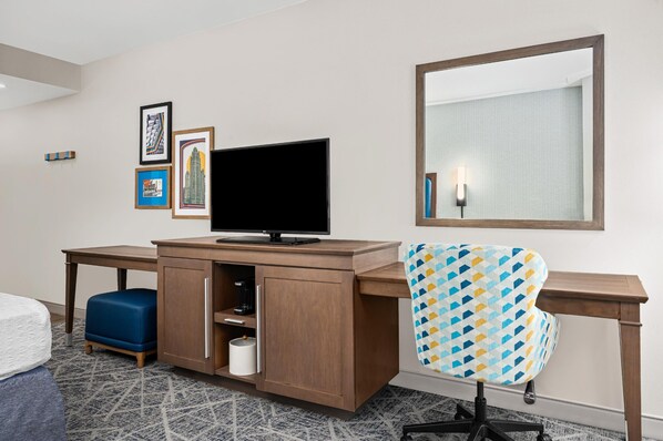 Une chambre d’hôtel avec un bureau en bois, une télévision, une chaise à motifs, un lit et un miroir.