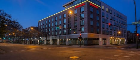 A modern hotel with a prominent 'Hampton Inn' sign, a corner location with a street view, and a crosswalk in front.