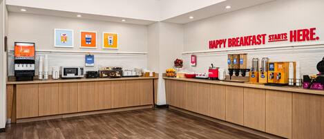 A clean, modern breakfast area with wooden counters, a juice dispenser, and a sign that reads 'HAPPY BREAKFAST STARTS HERE.'