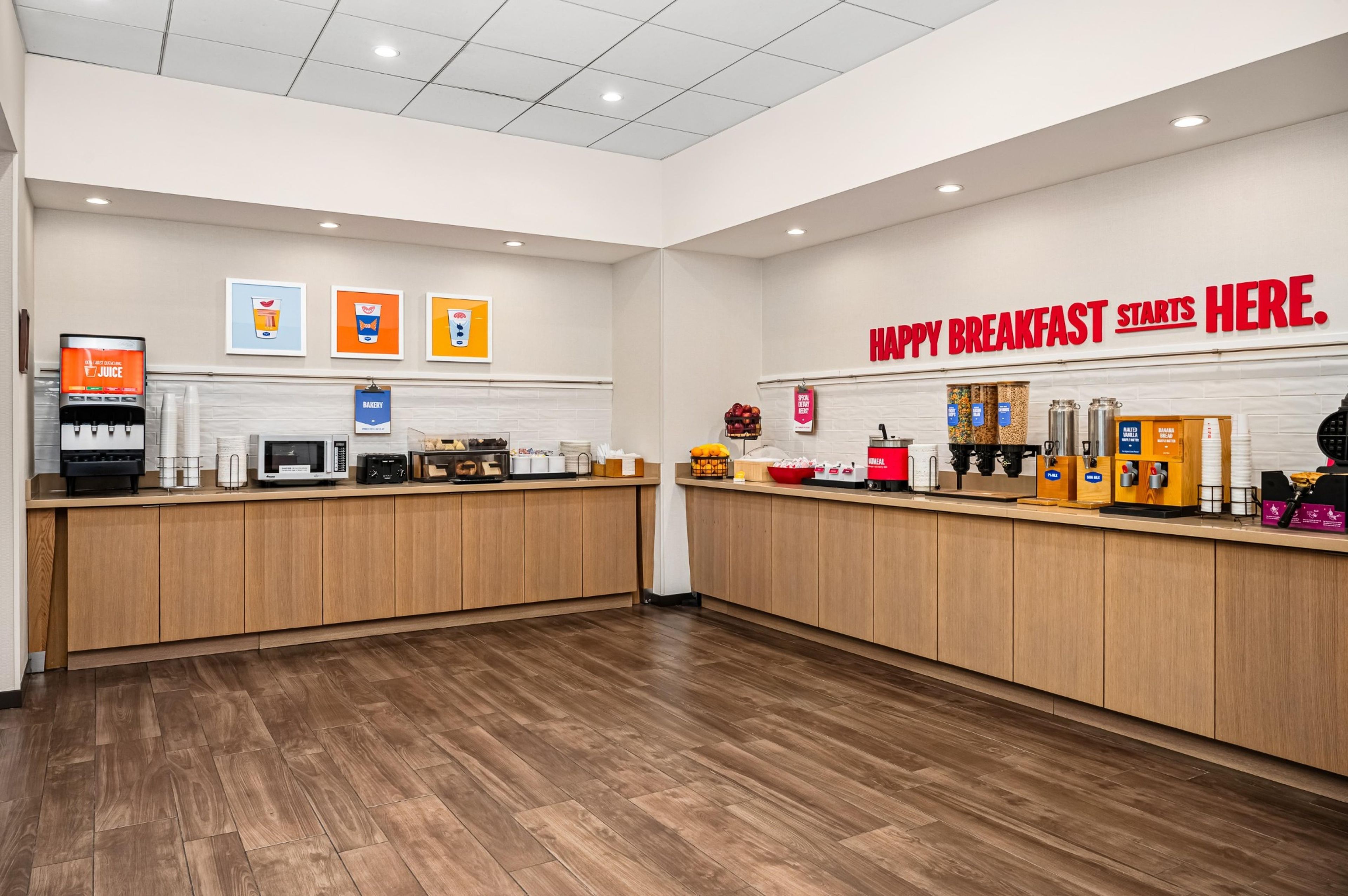 Un espace petit-déjeuner propre et moderne, avec des comptoirs en bois, un distributeur de jus et un panneau sur lequel on peut lire : « C’EST ICI QUE COMMENCE UN PETIT-DÉJEUNER RÉUSSI ».