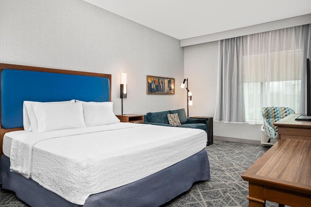 Hotelkamer met een groot bed, een bureau, een stoel, een nachtkastje, een lamp, een schilderij en een raam met gordijnen.