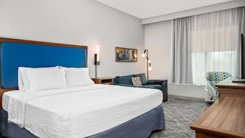 Une chambre d’hôtel avec un grand lit, un bureau, une chaise, une table de chevet, une lampe, un tableau et une fenêtre avec des rideaux.