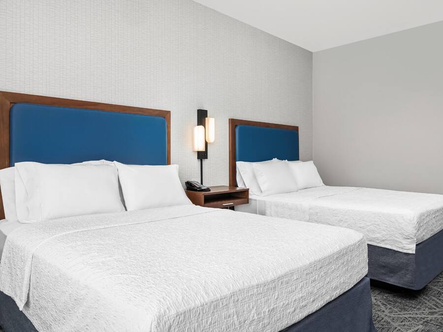 Habitación de hotel con dos camas, una mesita de noche con lámpara y una alfombra estampada.