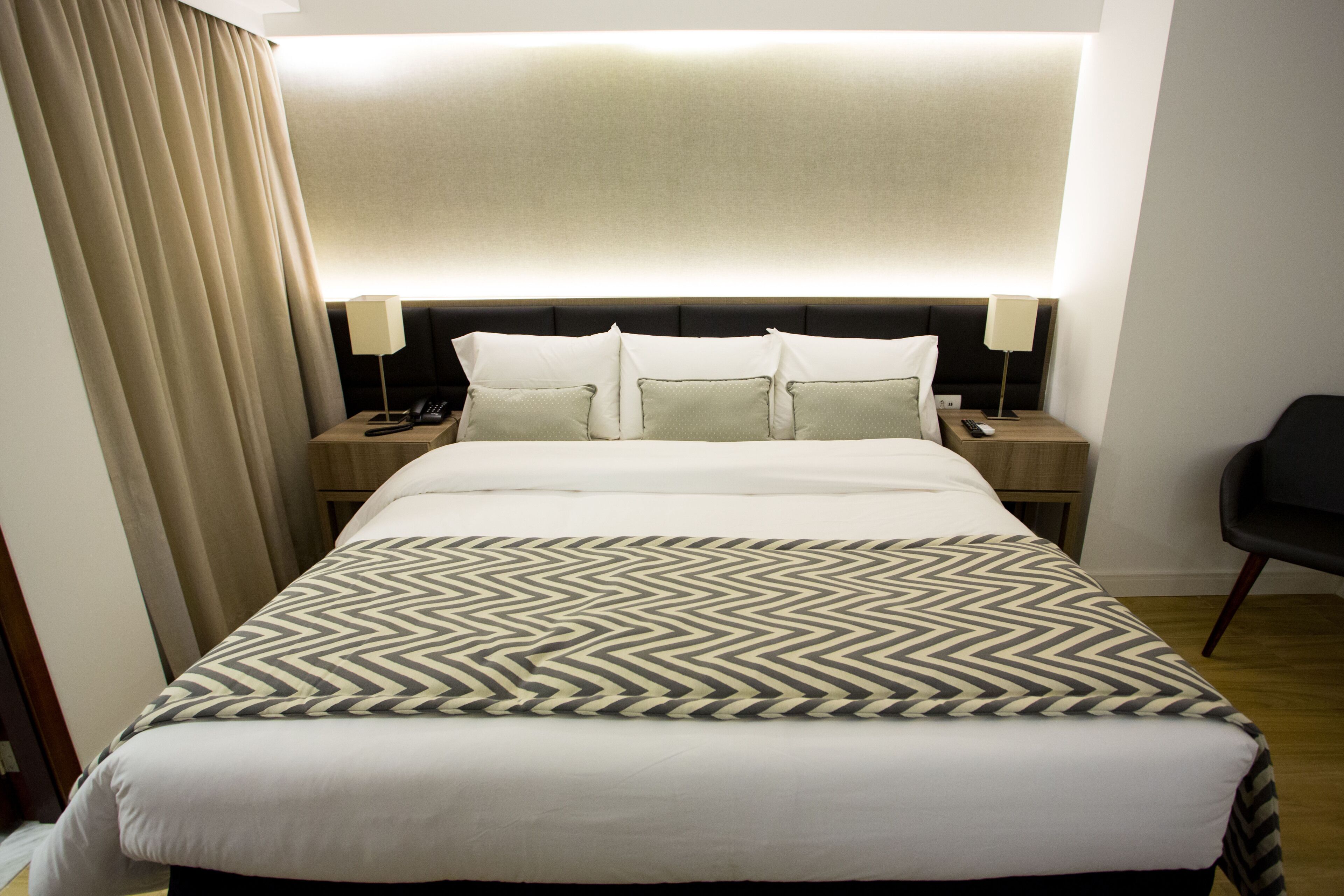A hotel room with a large bed, a headboard with integrated lighting, a bedside table with a lamp, and a chair.