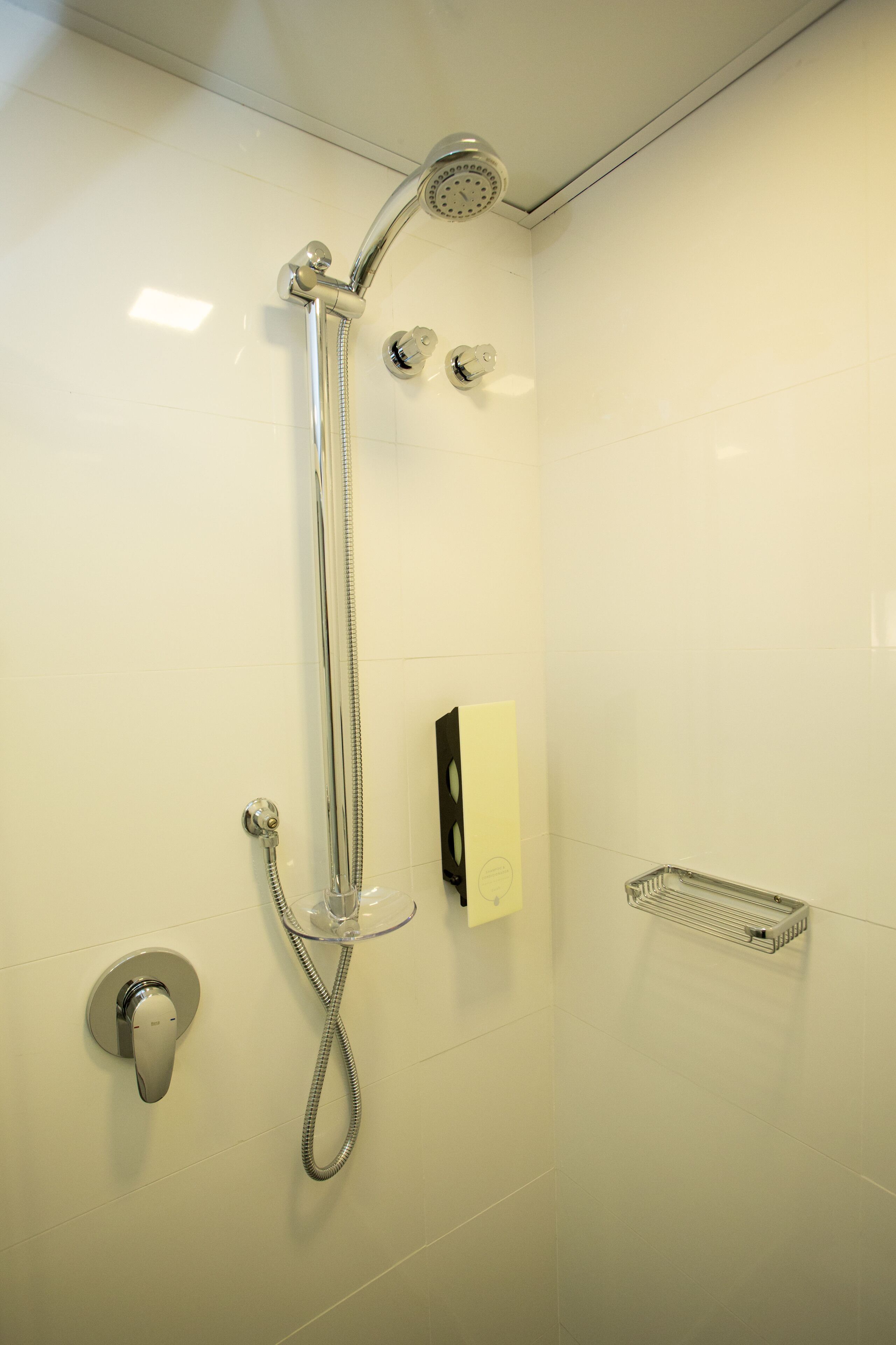 Eine Dusche mit Handbrause, Armaturen an der Wand und einem durchsichtigen Plastikregal.
