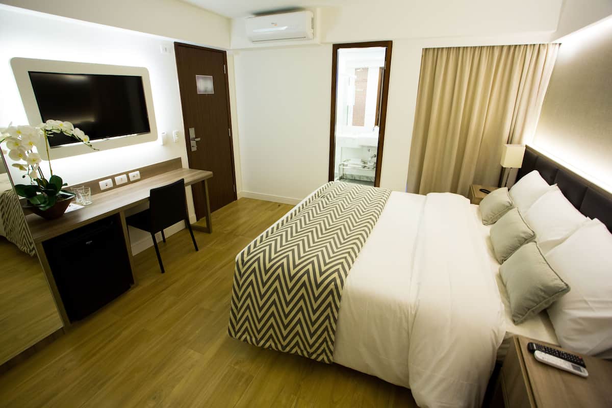 A hotel room with a bed, a desk, a chair, a TV, and a bathroom visible through an open door.