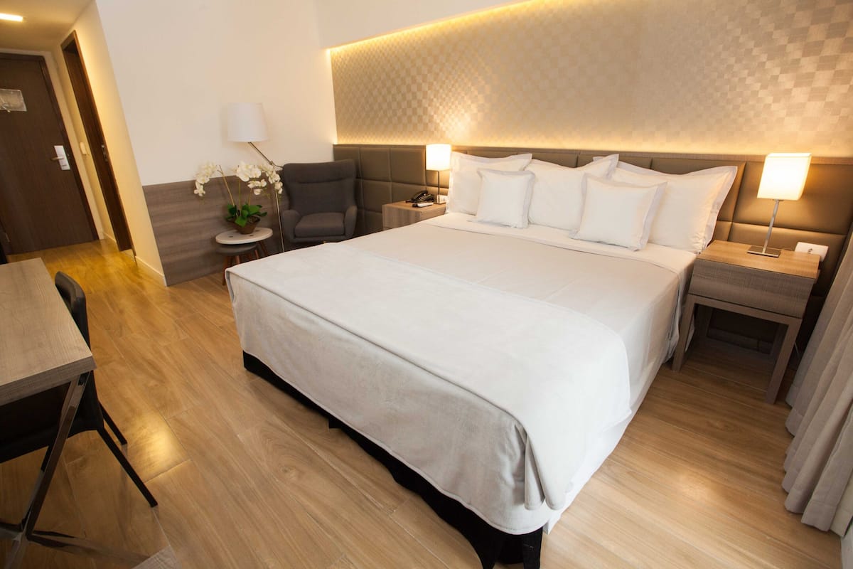 A hotel room with a large bed, two bedside tables, a desk, a chair, and a small table with a lamp and orchid plant.