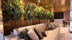 Un hall d’hôtel moderne avec un canapé en cuir blanc, plusieurs coussins à motifs et une rangée d’arbres artificiels devant un mur en bois.