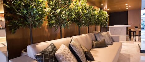 A modern hotel lobby with a white leather sofa, multiple patterned cushions, and a row of artificial trees against a wooden wall.