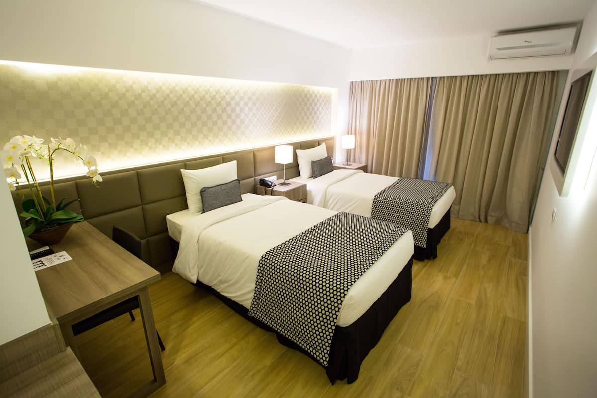 Ein Hotelzimmer mit zwei Betten, einem Schreibtisch, einer Zimmerpflanze und einem großen Fenster mit Vorhängen.