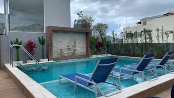 A swimming pool with lounge chairs, a stone wall, and a modern building in the background.