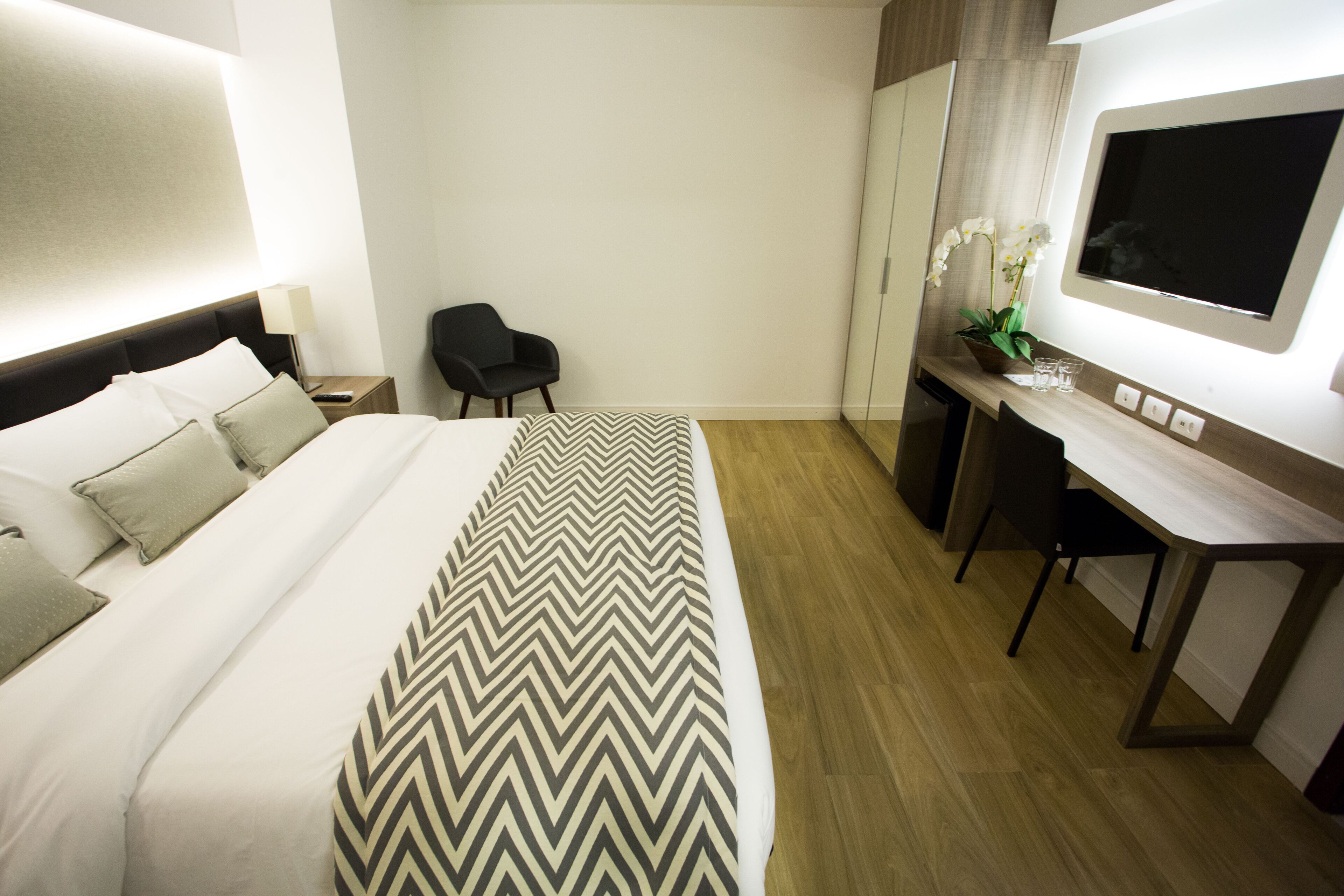 Quarto de hotel com uma cama grande, uma escrivaninha, uma cadeira, uma televisão e uma pequena mesa com uma planta.