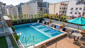 Une piscine sur le toit, agrémentée de chaises longues et d’une table, entourée de plantes en pot et offrant une vue sur les bâtiments de la ville.