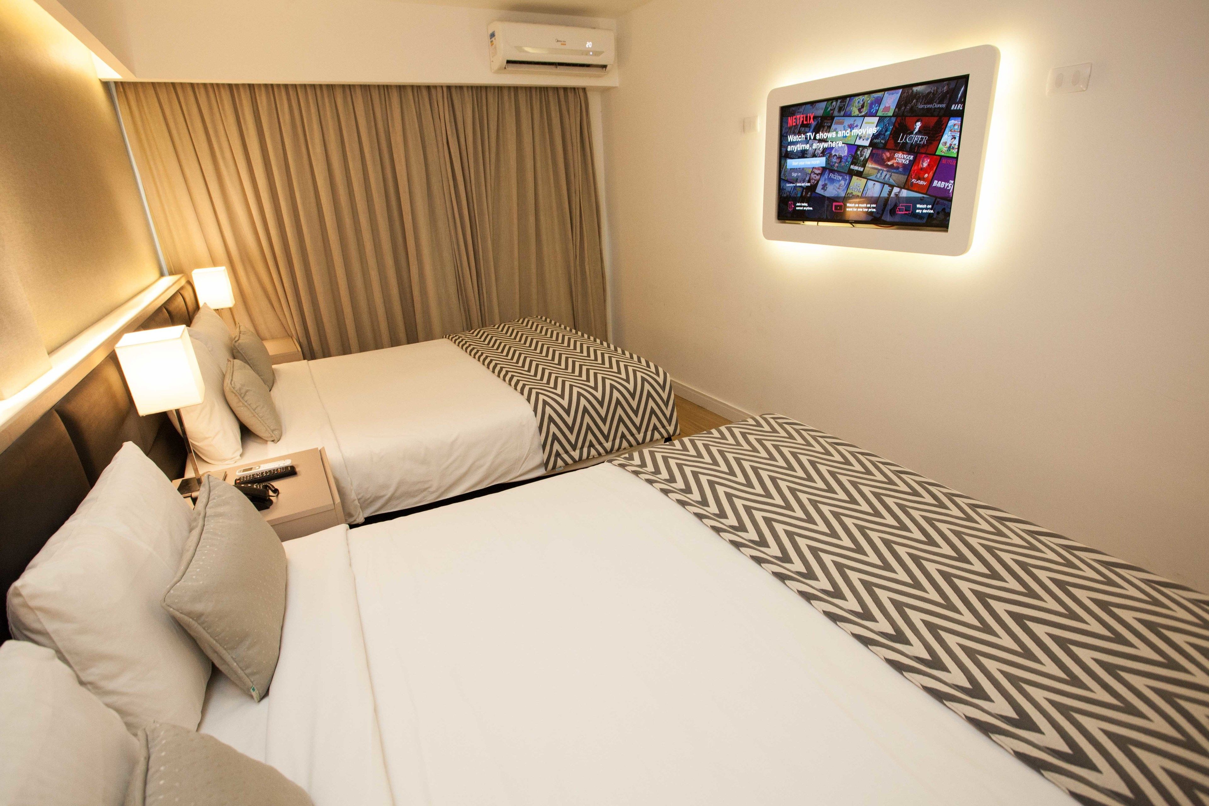 A hotel room with two beds, a TV mounted on the wall, and an air conditioning unit above the beds.