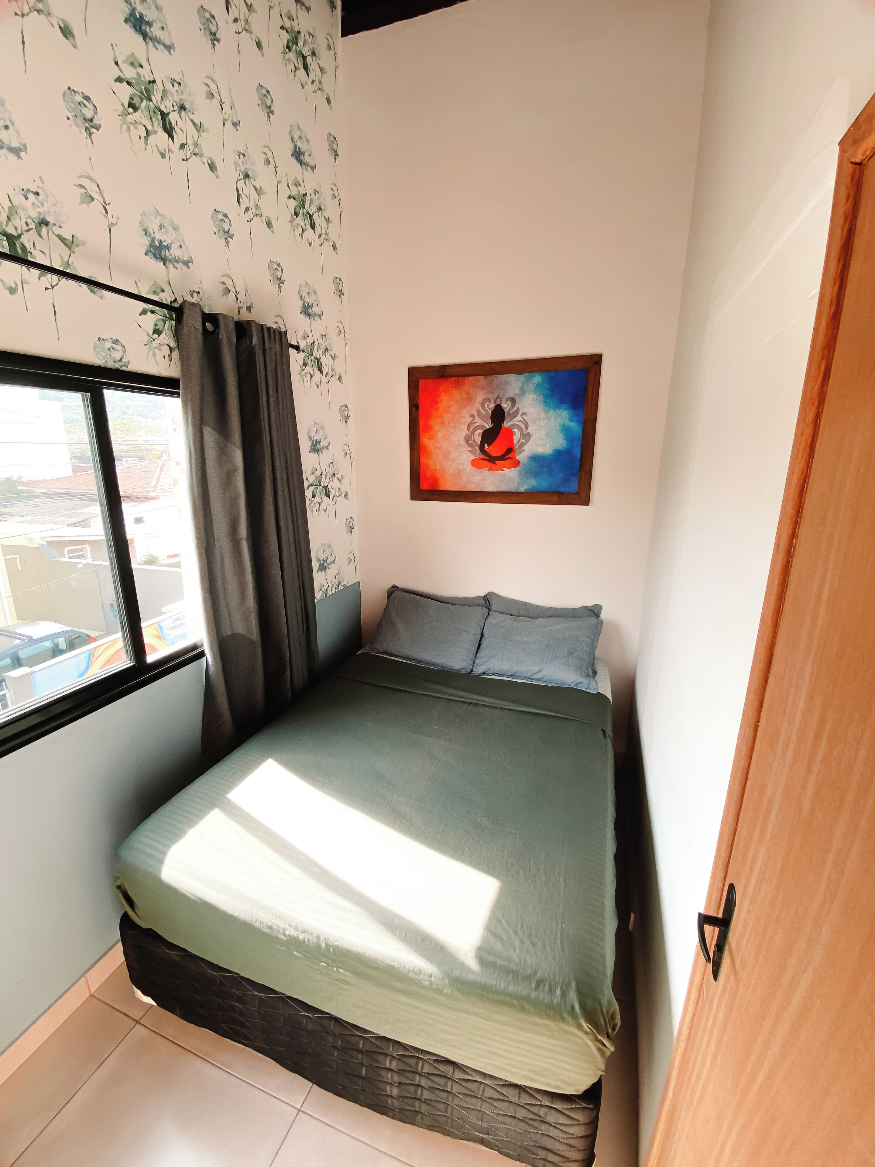 Un dormitorio pequeño con una cama, una ventana, un papel tapiz floral en la pared y un cuadro.