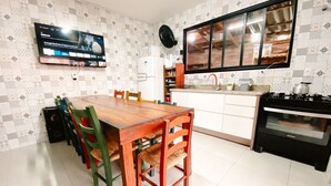 A kitchen with a wooden dining table, chairs, a TV mounted on the wall, a refrigerator, a stove, and a window with a view of the kitchen. - Sagui Hostel (Florianópolis)