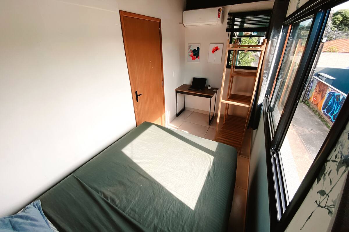 A room with a bed, a desk with a laptop, and a window with a view of the outside.