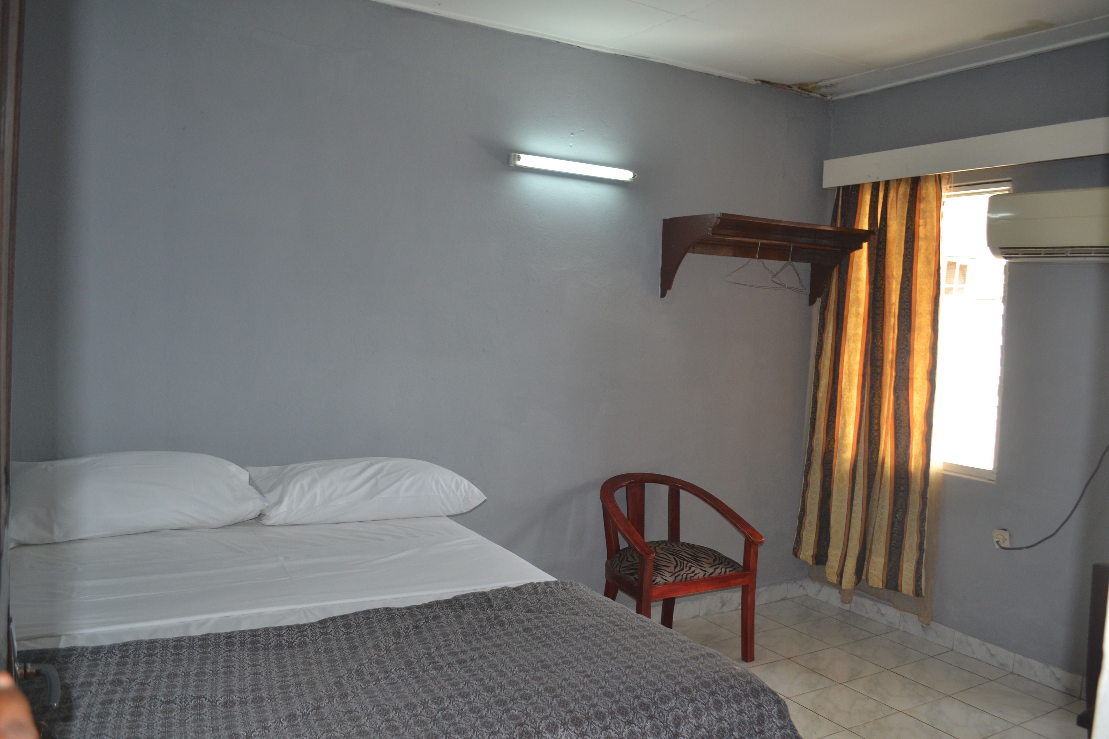 Una habitació amb un llit, una cadira, una prestatgeria penjada a la paret i una finestra amb cortines.