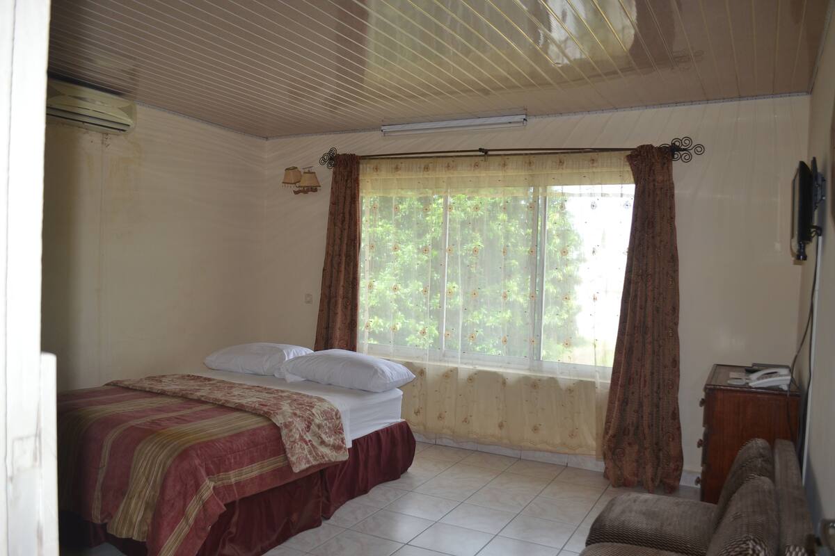 A bedroom with a bed, a window with curtains, a radiator, and a small wooden side table.