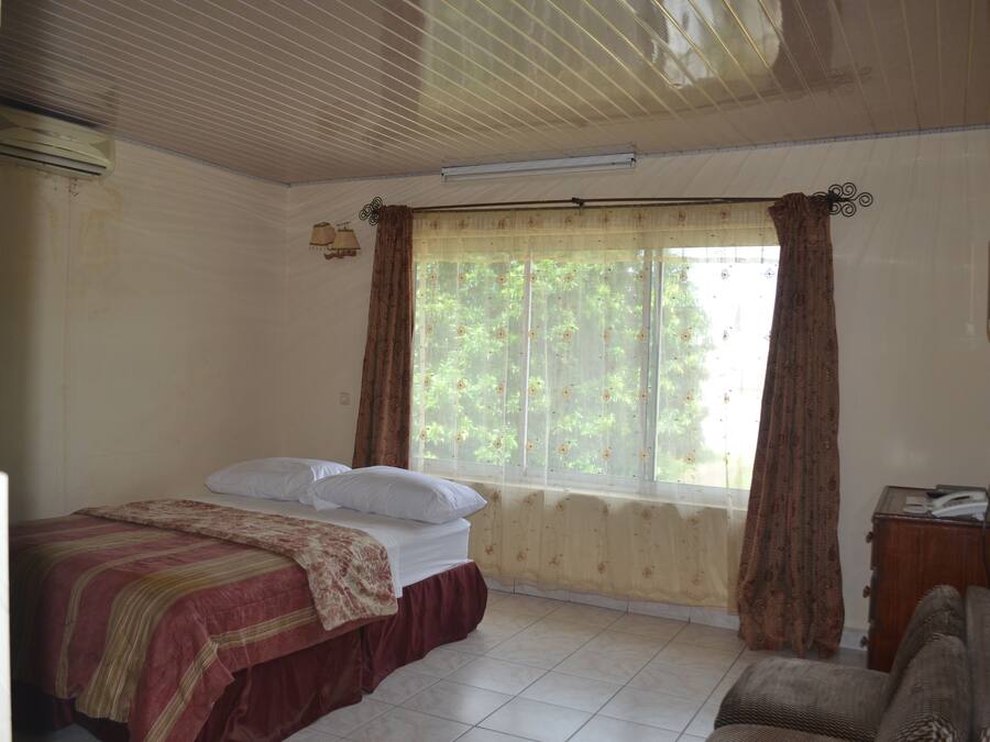 A bedroom with a bed, a window with curtains, a radiator, and a small wooden side table.