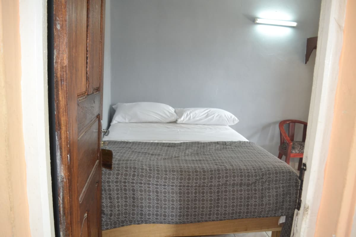 A small, narrow room with a single bed, a wooden door, and a chair.