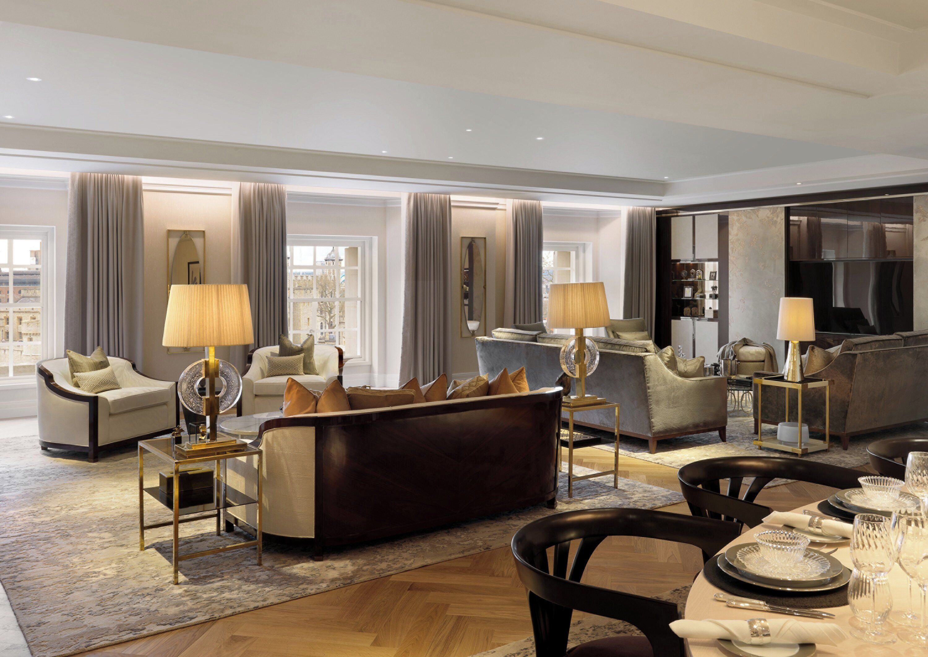 A spacious living room with a dark brown sofa, beige armchairs, and a blue sofa. There are two lamps on side tables, a coffee table, and a dining area with a set table.