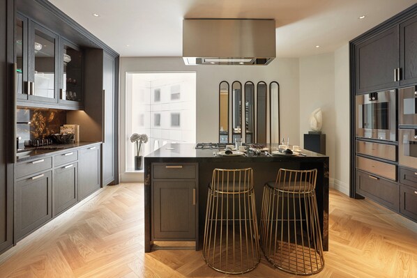 A modern kitchen with dark cabinetry, a central island with stools, and a large window providing natural light. - Four Seasons Hotel London at Tower Bridge (London)