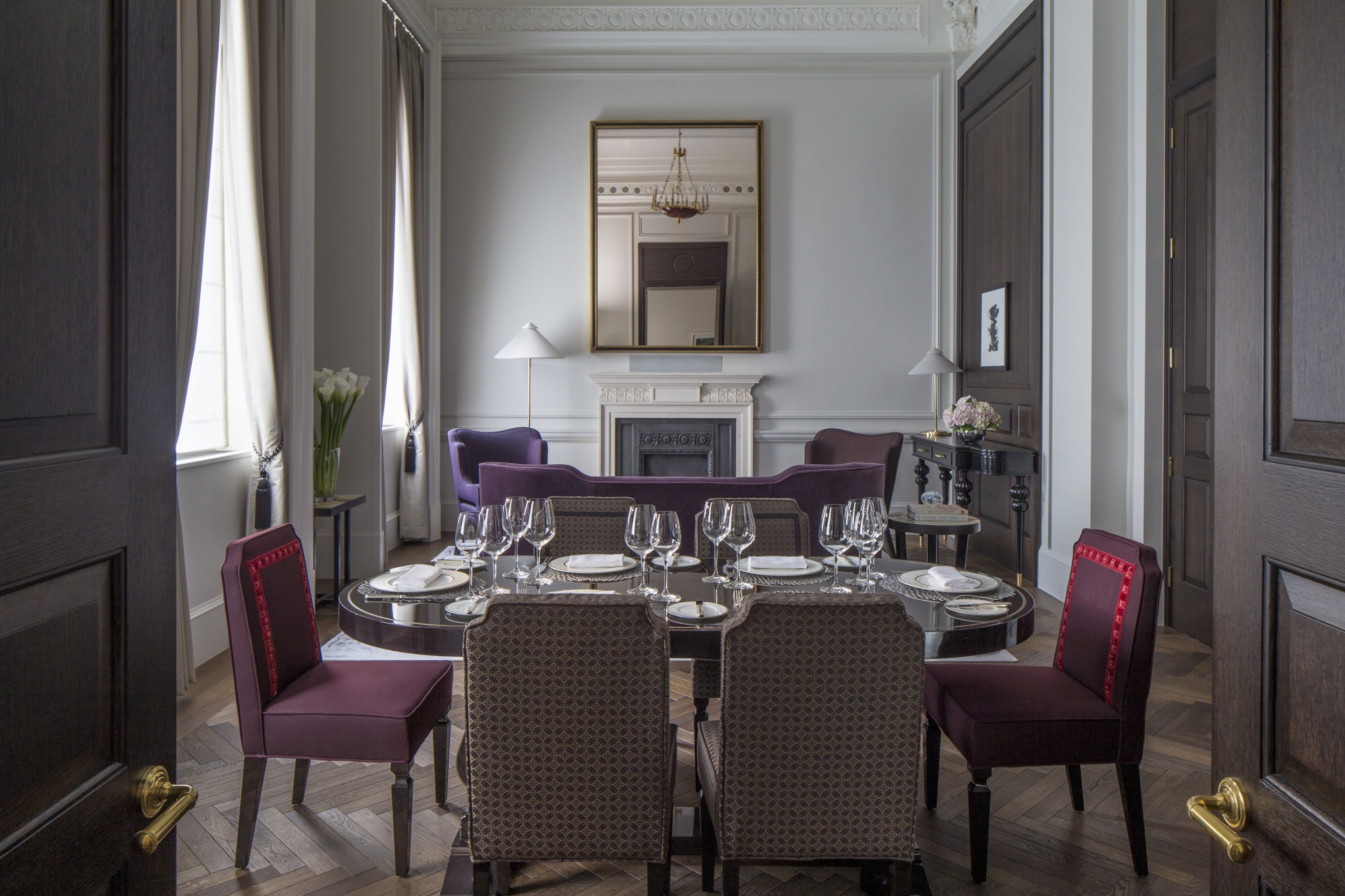 A formal dining room with a large table set for six, flanked by two purple chairs with red detailing. A fireplace with a mirror above it is visible in the background.
