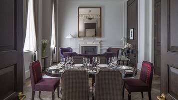 A formal dining room with a large table set for six, flanked by two purple chairs with red detailing. A fireplace with a mirror above it is visible in the background.