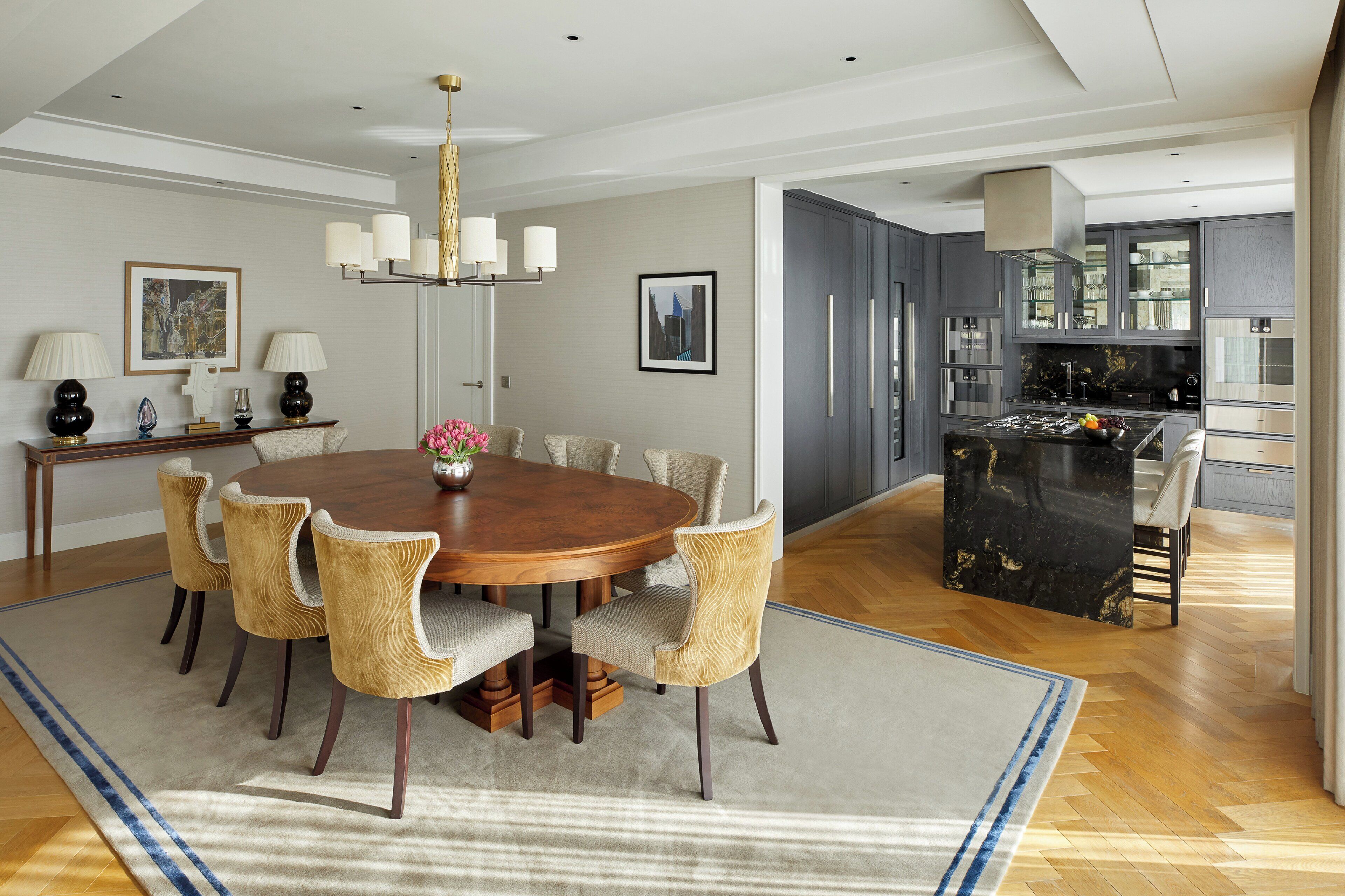 A modern dining area with a wooden table and chairs, a chandelier, and a kitchen in the background.