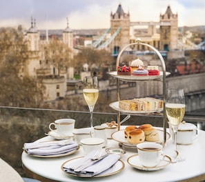 A three-tiered dessert stand with various pastries and a glass of champagne on a table with tea cups and saucers, set against a cityscape with a bridge and historic buildings. - Four Seasons Hotel London at Tower Bridge (London)