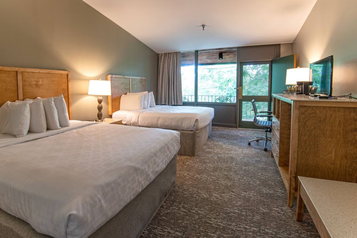 Une chambre d’hôtel avec deux lits, un bureau, une chaise et une grande fenêtre donnant sur des arbres.
