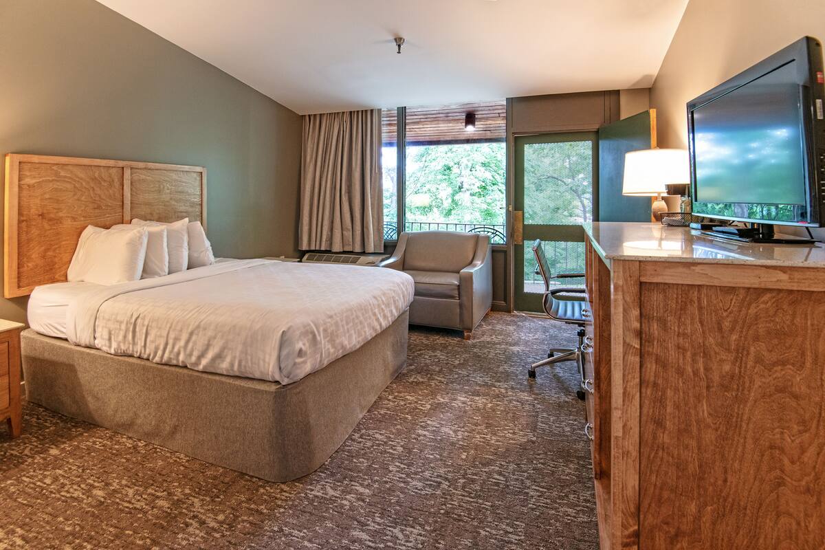 Une chambre d’hôtel avec un grand lit, un bureau avec une télévision, une chaise et une vue sur la verdure environnante.