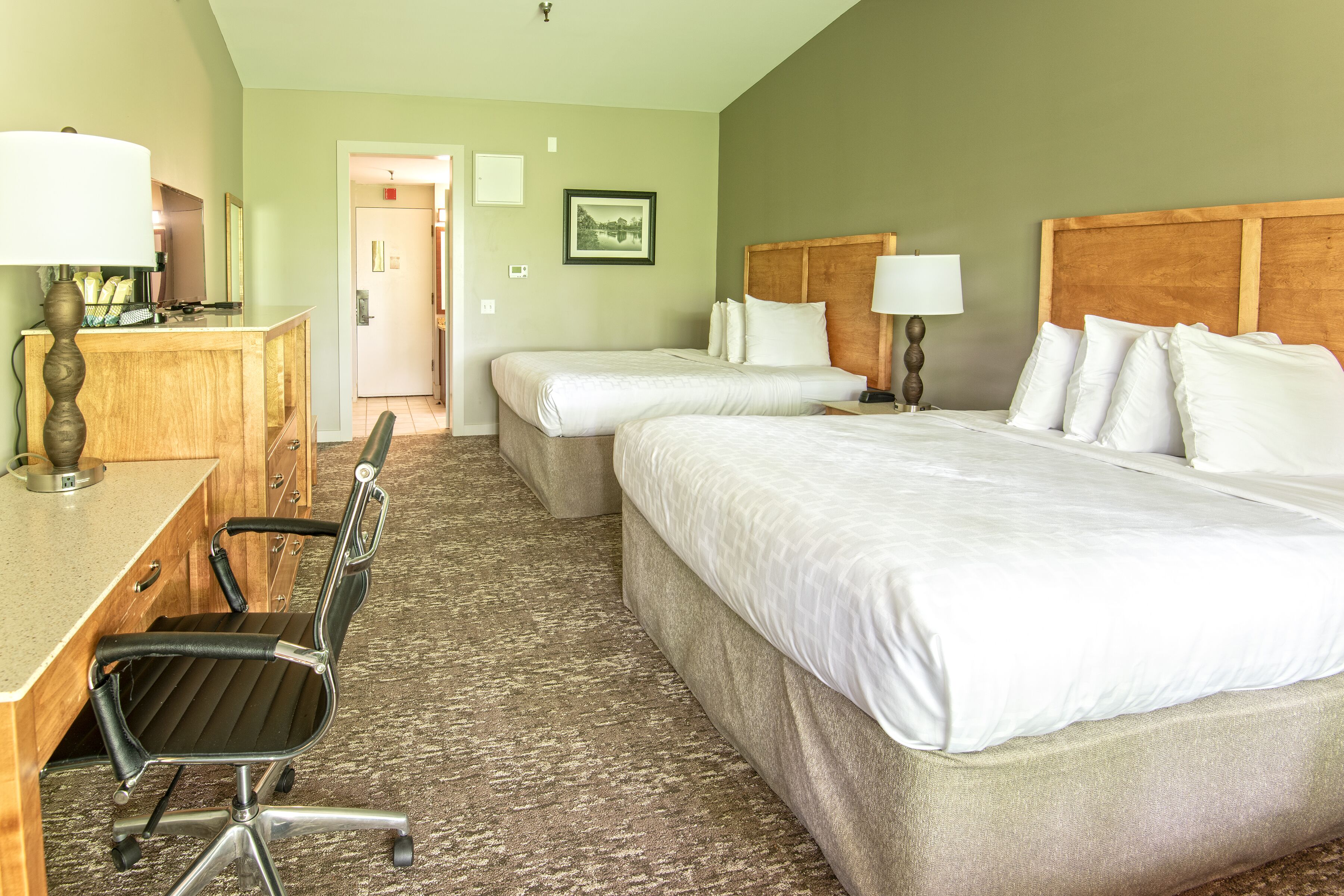 A hotel room with two beds, a desk, a chair, and a lamp.