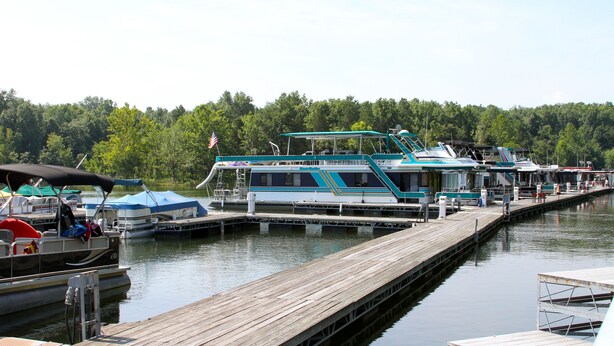 A dock with several boats, including a large houseboat with a flag, docked at a marina surrounded by trees. - Rough River Dam State Resort Park (Falls of Rough)