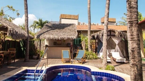 A swimming pool surrounded by palm trees and thatched-roof structures, with a clear blue sky in the background.