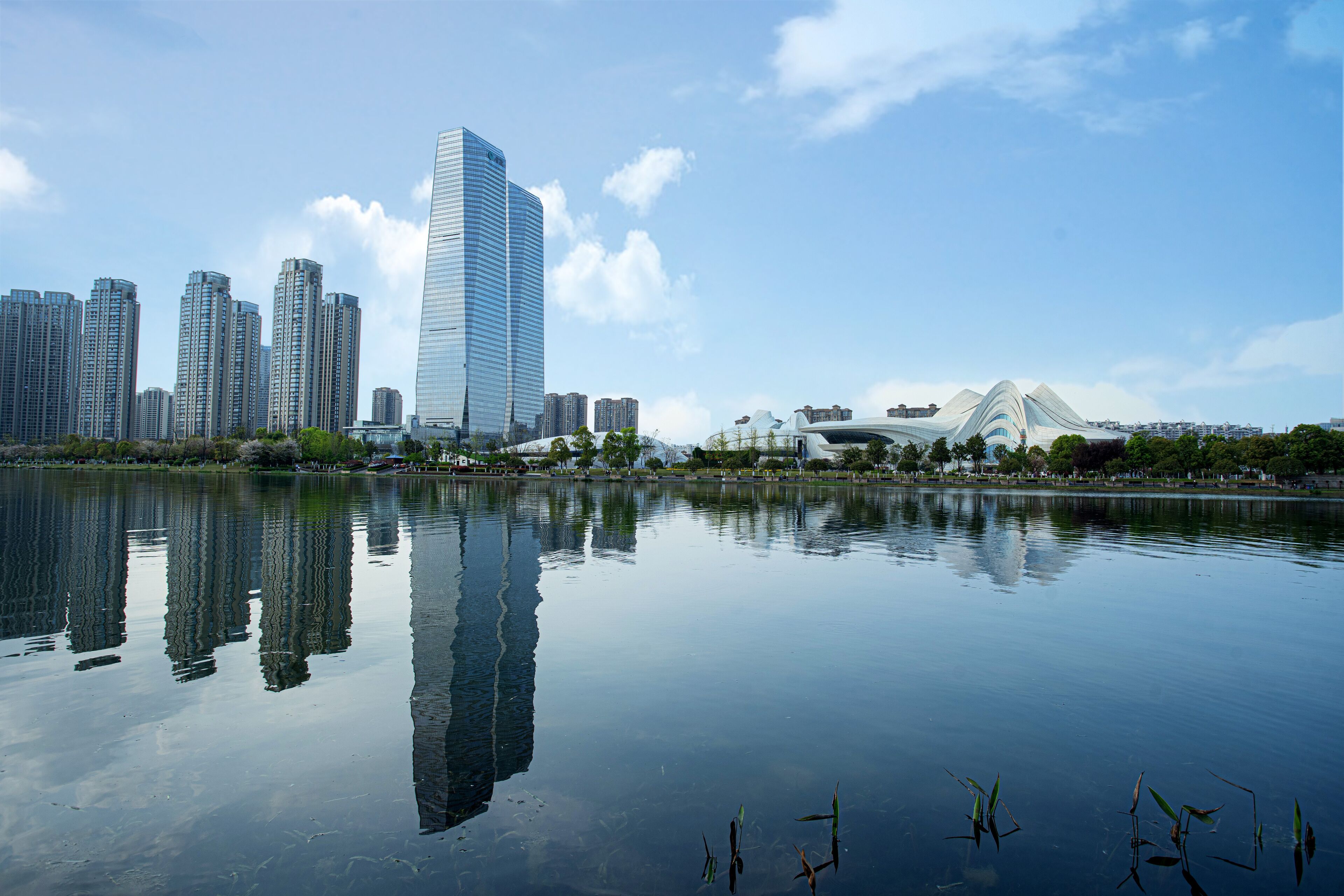 Un paysage urbain avec de hauts immeubles et une structure architecturale moderne, le tout surplombant une étendue d’eau calme.