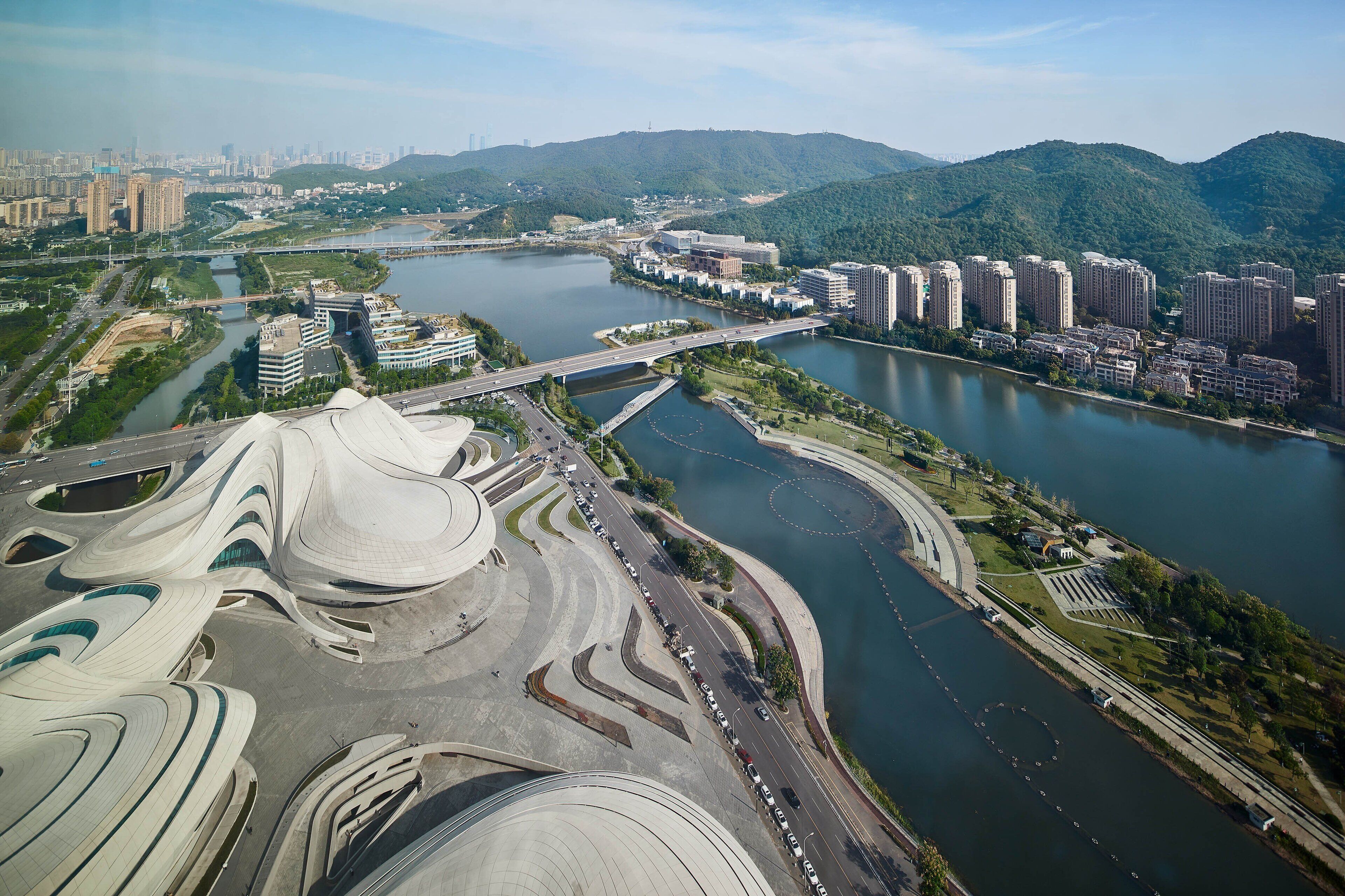 Vue aérienne d’un quartier urbain moderne, avec une rivière, des ponts et un bâtiment remarquable à l’architecture particulière, caractérisé par des toits incurvés.