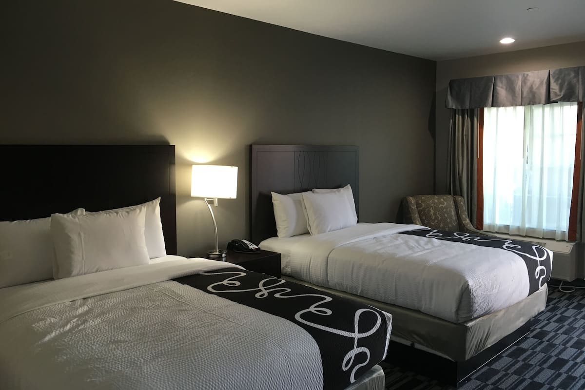 Une chambre d’hôtel avec deux lits, une table de chevet, une lampe, une chaise et une fenêtre avec des rideaux.