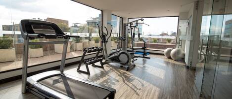 A modern gym with a treadmill, elliptical, and weight machines, large windows with a city view, and a wooden floor.
